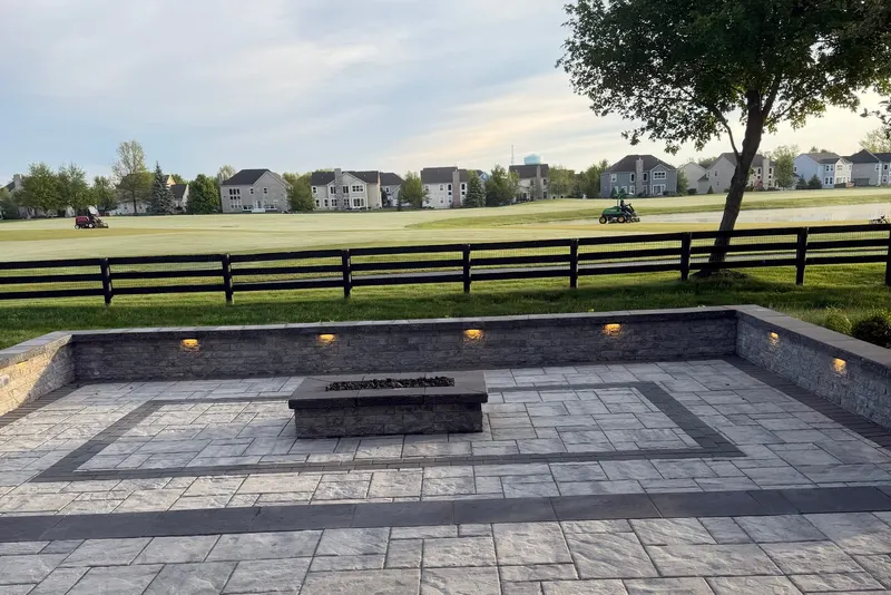 Large stamped-stone patio with a contrasting border, low stone seating wall with built-in lighting, and a gas fire table, overlooking open green space at dusk